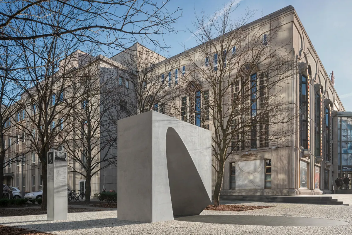A pale concrete block with a smooth curved hollow, standing among trees on a paved square beside a large stone building