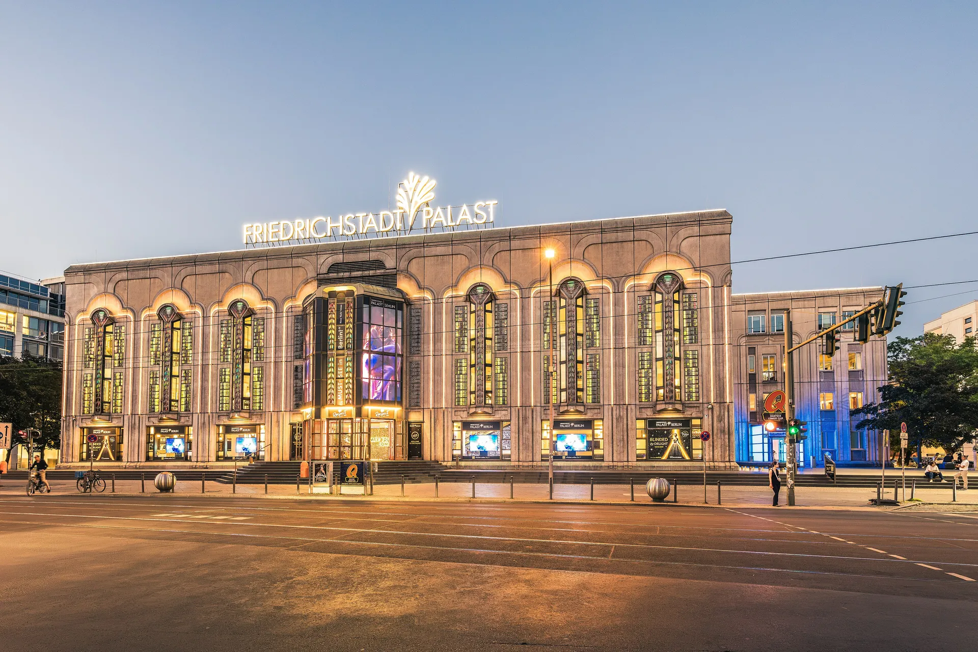 The lit front of the Friedrichstadt-Palast on Friedrichstraße at dusk, a wide stone building with tall arched windows, a glass tower entrance and a glowing sign on the roof